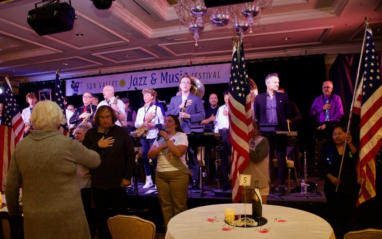 The festival's Marching Band on stage under the Sun Valley Jazz and Music Festival banner, with local Girl Scouts presenting the colors, in a photograph from the festival site. The site says the band is led by conductor Tom Dale.